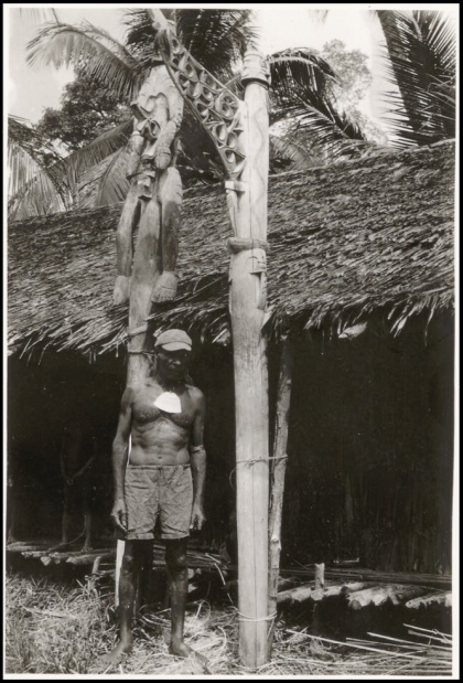 Un homme debout devant une maison en paille, tenant une lance, avec un toit de palme et une sculpture en bois au-dessus de lui.
