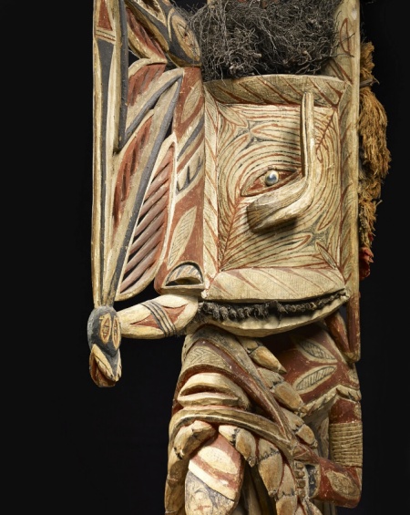 Une sculpture en bois décorée, représentant un visage stylisé avec des motifs géométriques et des couleurs vives, ornée de matière végétale sur le dessus.