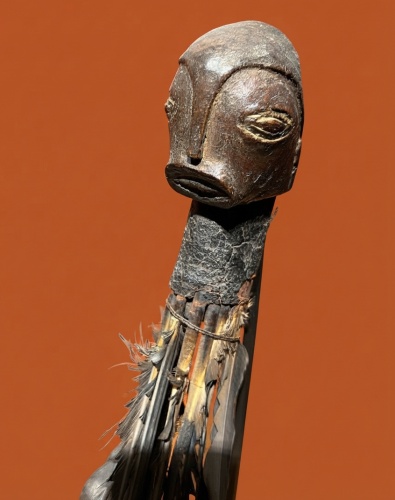 Une sculpture en bois représentant une tête stylisée sur un long corps décoré de plumes, sur fond orange.