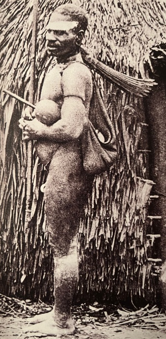 Un homme debout en tenue traditionnelle, tenant un gourde et un bâton, devant une structure en paille.